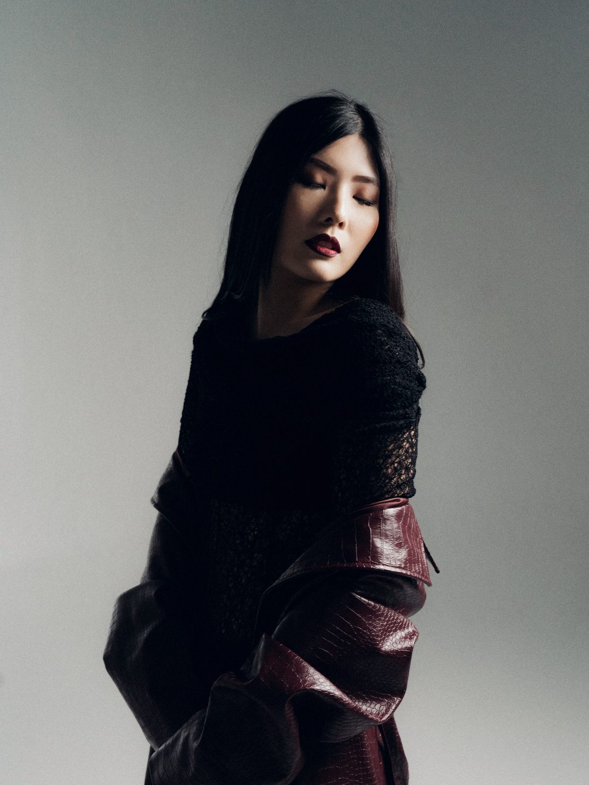 Retrato de estudio sobre fondo gris: modelo de perfil con blusa negra y falda de cuero burdeos, luz lateral suave.