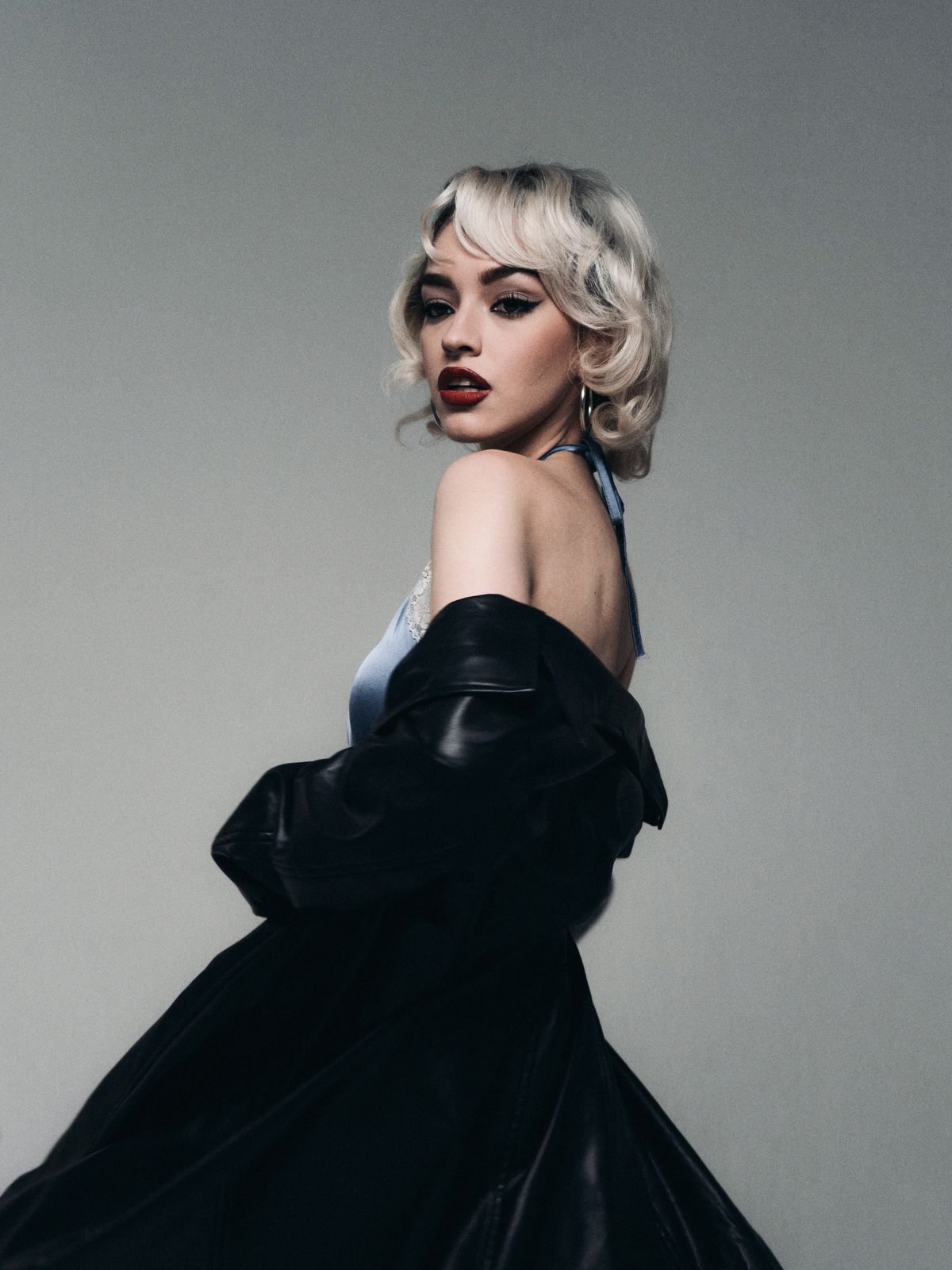 Luna Vesper, Chapter 01: la modelo de perfil sobre fondo gris de estudio, con abrigo negro de piel caído sobre los hombros y vestido de raso azul claro